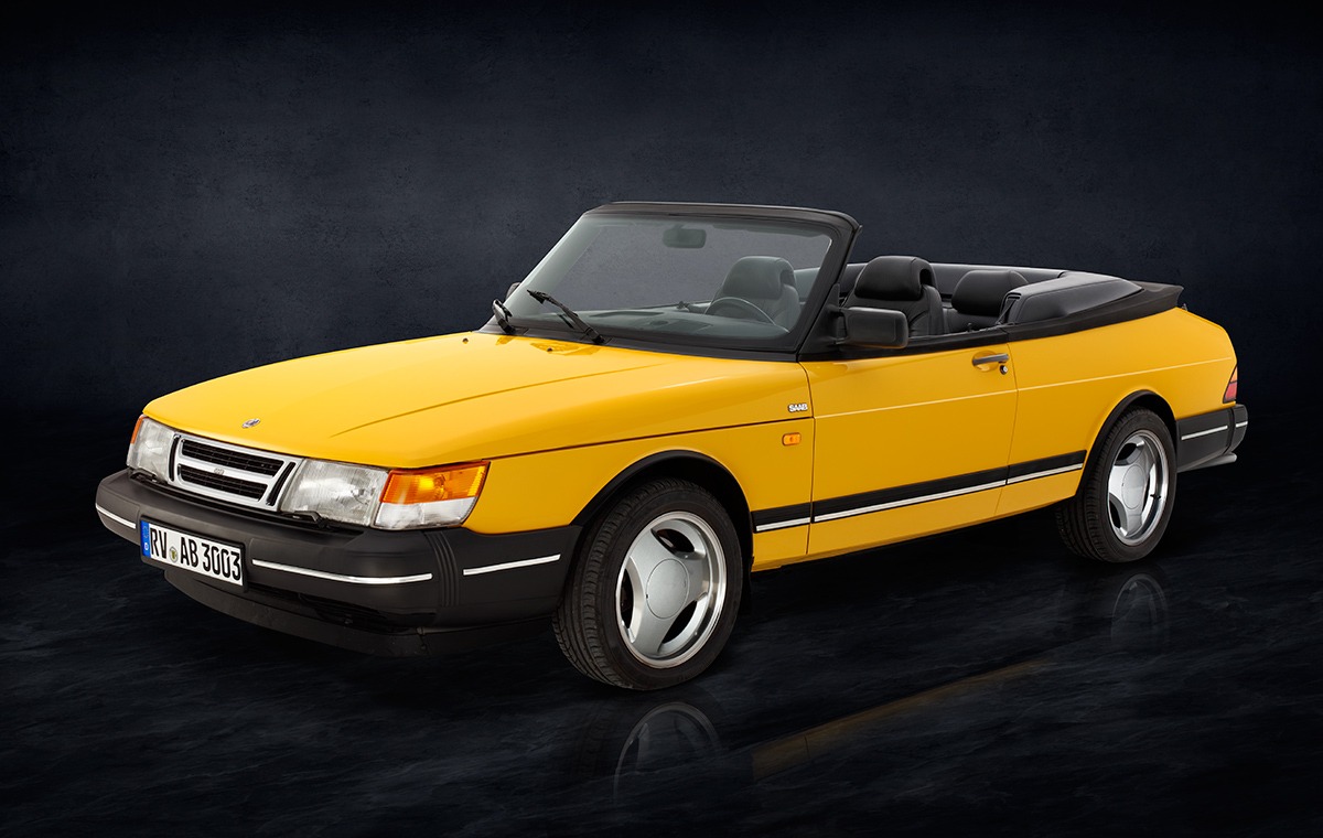 Saab 900 i Cabriolet Front Ein gelber Sportwagen-Cabrio, Modell Saab, steht seitlich auf einer dunklen, einfarbigen Oberfläche. Das Auto hat schwarze Details und ein offenes Verdeck, sichtbar sind die beiden vorderen Sitze im Innenraum.