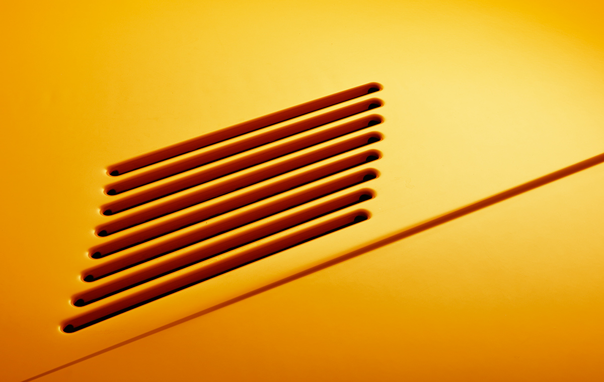 Saab 900 i Cabriolet Lufteinlass Detailaufnahme von einer orangefarbenen Oberfläche mit mehreren parallel angeordneten Schlitzöffnungen, die Schatten werfen.