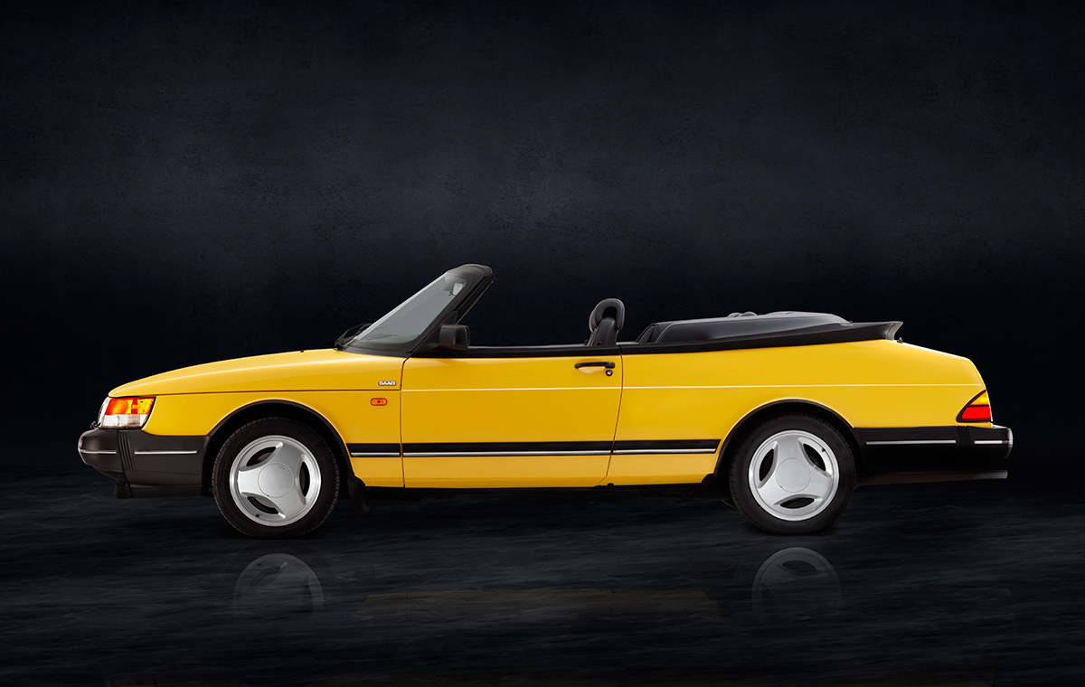 Saab 900 i Cabriolet Seite Ein gelber Cabriolet mit schwarzem Verdeck, seitlich abgebildet. Das Auto hat eine sportliche Silhouette und auffällige weiße Felgen. Der Hintergrund ist dunkel und hebt das Fahrzeug hervor.