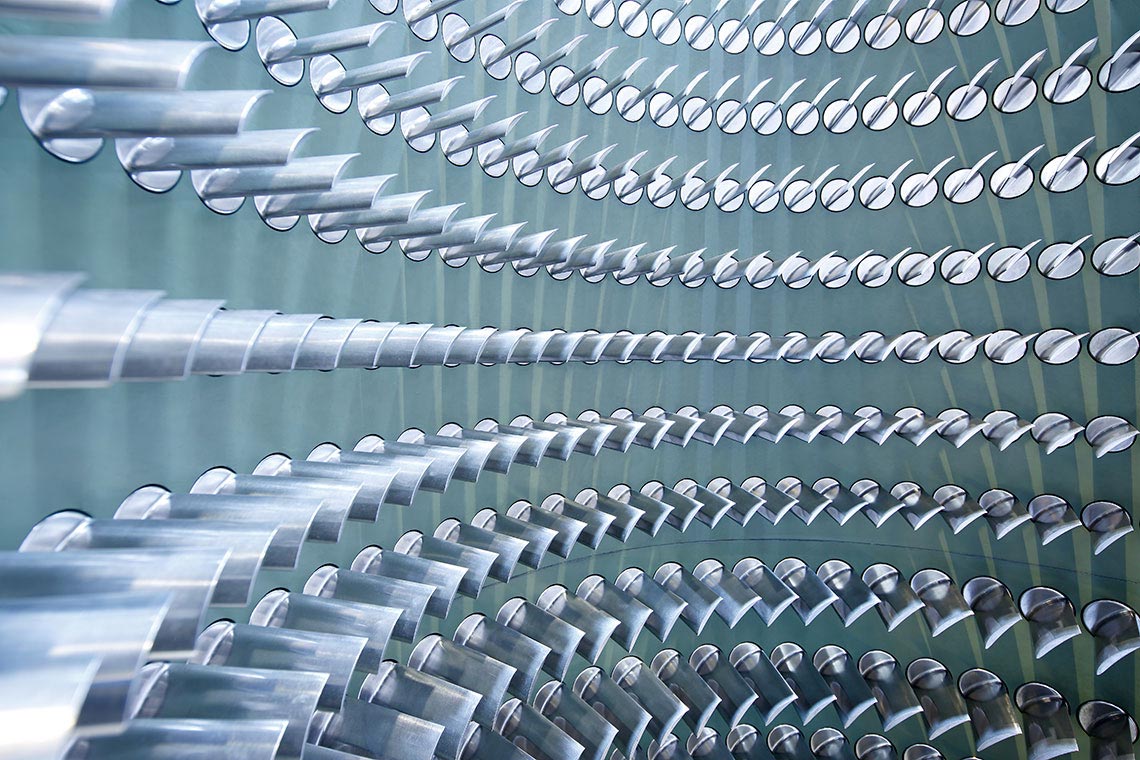 Schaufeltraeger Eine abstrakte Nahaufnahme von glatten, metallischen Rohren, die in geschwungenen, parallel angeordneten Reihen angeordnet sind. Die Rohre reflektieren Licht und erzeugen eine dynamische, wellenartige Struktur. Der Hintergrund ist in sanften, gedeckten Farben gehalten.