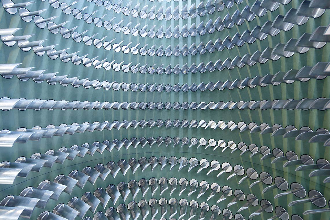 Schaufeltraeger Ein Muster von vielen silbernen Rohren, die kreisförmig angeordnet sind. Die Rohre sind teilweise spiegelnd und reflektieren das Licht, was einen dreidimensionalen Effekt erzeugt. Der Hintergrund ist in einem sanften Grünton gehalten.