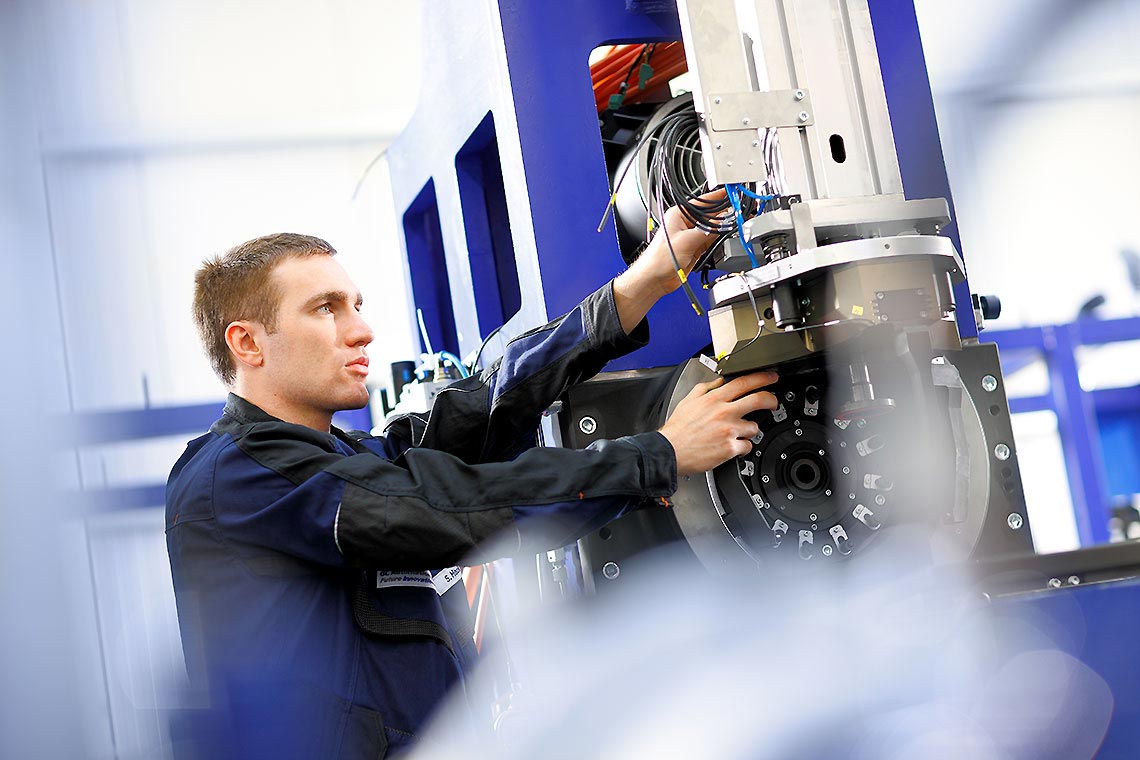 Montageszene Fertigungsanlage Ein Techniker arbeitet an einer Maschine in einer Industrieumgebung. Er hat eine konzentrierte Miene und hält eine Hand an einem Teil der Maschine, während er es anpasst. Die Umgebung ist modern und technisch gestaltet.