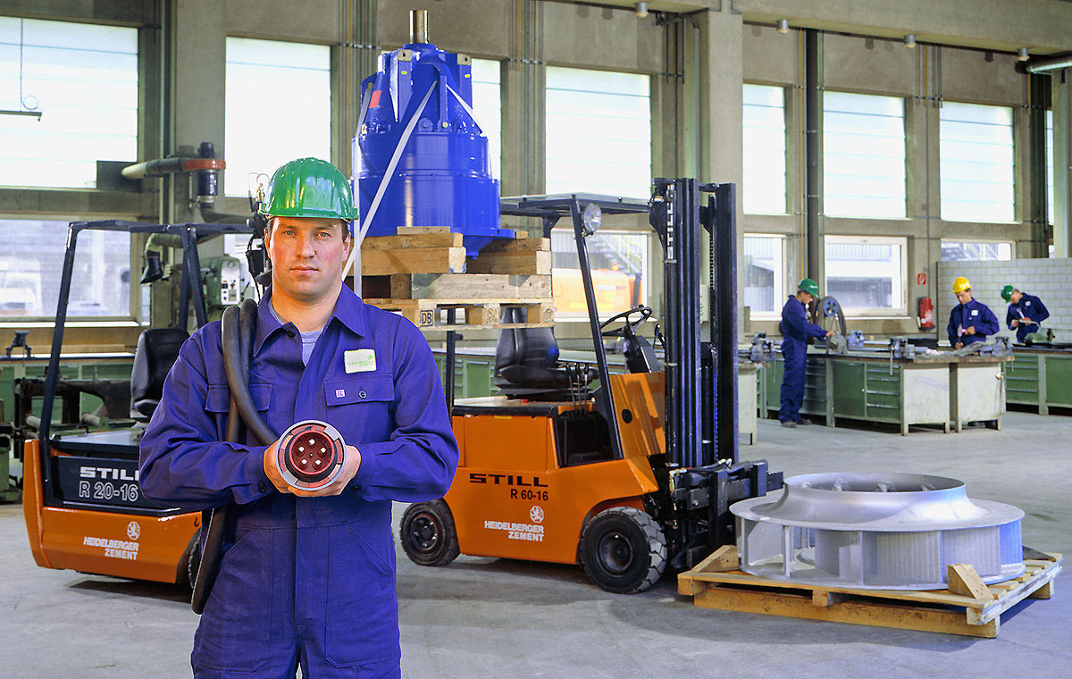 Schlosserei Ein Arbeiter in blauer Arbeitskleidung und mit einem grünen Helm steht in einer Industriehalle. Er hält ein Werkzeug in der Hand. Im Hintergrund sind Gabelstapler und weitere Arbeiter zu sehen, die an Maschinen arbeiten.