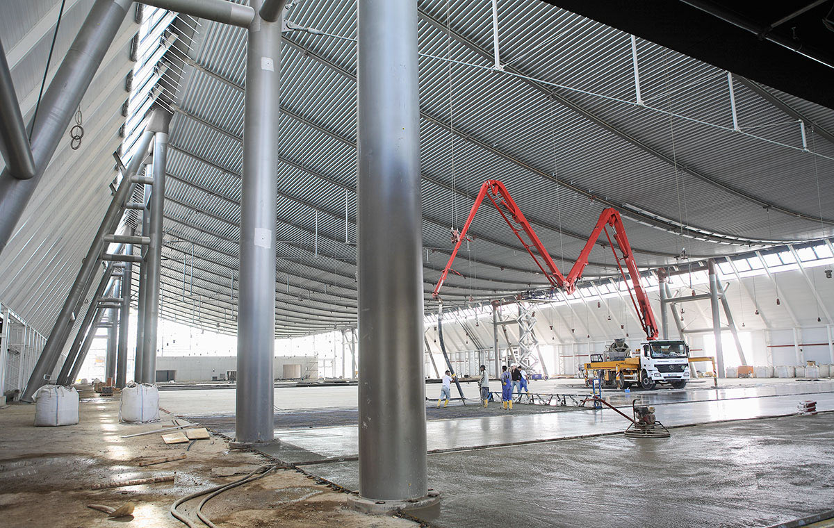 Baustelle Landesmesse Stuttgart Ein großer, leerer Innenraum mit hohen, metallic schimmernden Wänden und einem gewölbten Dach. In der Mitte befindet sich ein Betonmischer mit einem langen Schlauch, während Arbeiter auf dem Boden beschäftigt sind. Stützen aus Edelstahl sind sichtbar.