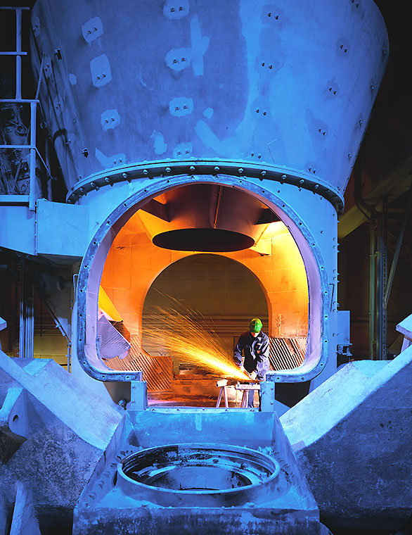 Instandhaltung Ein Arbeitsbereich in einer Industrieanlage, in dem ein Mitarbeiter mit Schutzkleidung an einem Maschineninneren arbeitet. Aus dem Inneren der Maschine strömt ein helles, orangefarbenes Licht, während Funken sprühen. Die Umgebung ist stark beleuchtet und metallisch geprägt.