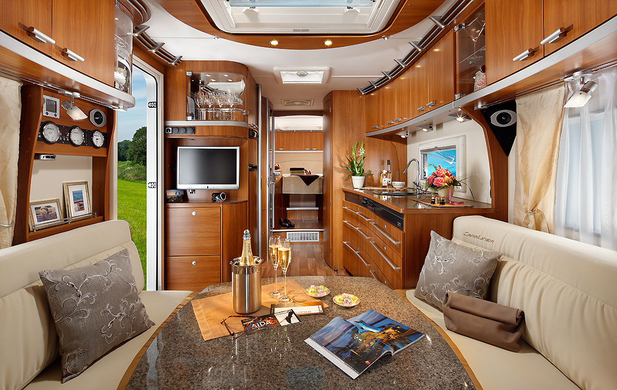 Ein modernes und elegantes Interieur eines Wohnmobils, das mit Holzverkleidungen, einem Esstisch und einer Sitzecke ausgestattet ist. Auf dem Tisch stehen Sektgläser, ein Kühlbehälter und Snacks. Im Hintergrund sieht man eine Küchenzeile und einen Fernseher. Große Fenster lassen Tageslicht herein.