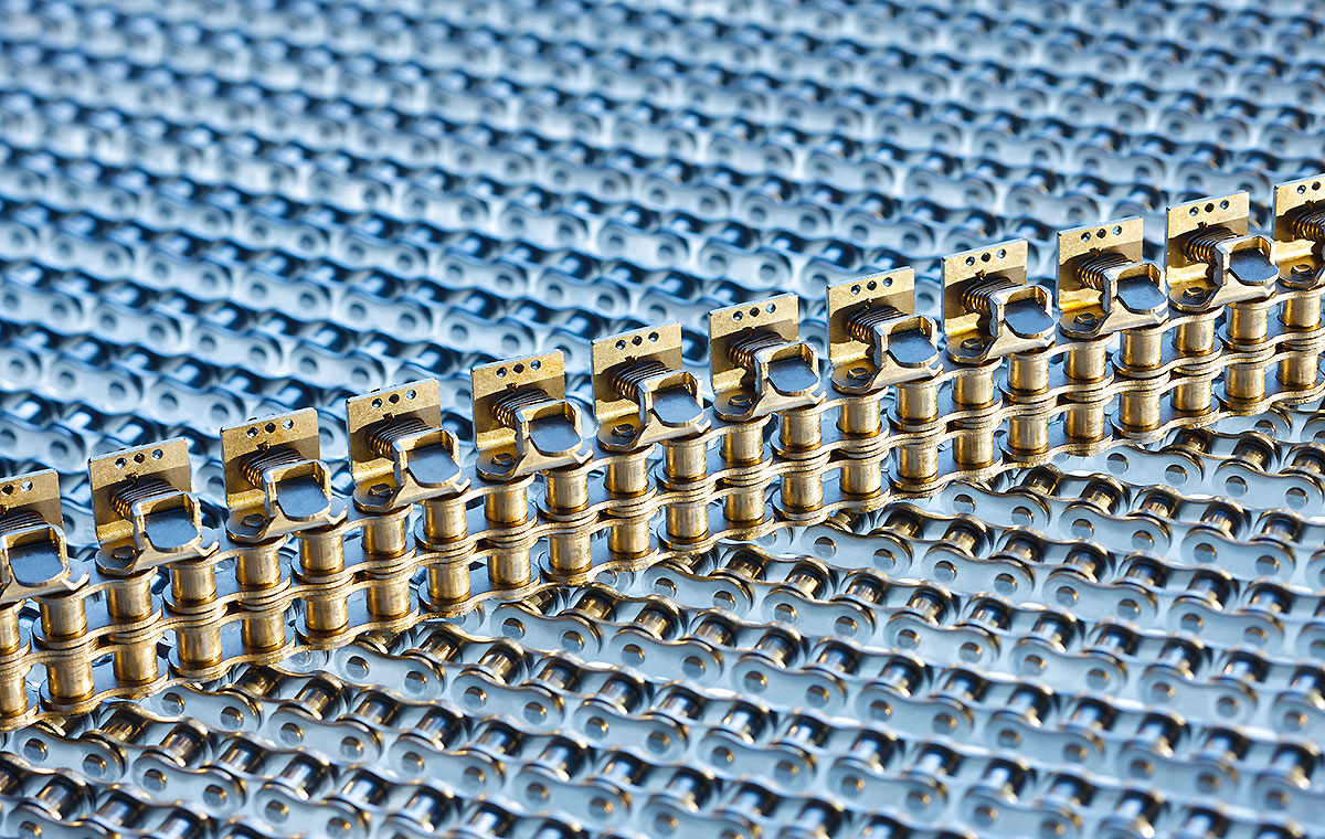 Eine Nahaufnahme von metallischen Kettenelementen auf einem blauen Hintergrund. Die Kette ist aus goldfarbenem Metall gefertigt und verläuft in gerader Linie. Die Struktur und Details der Kette sind gut sichtbar.