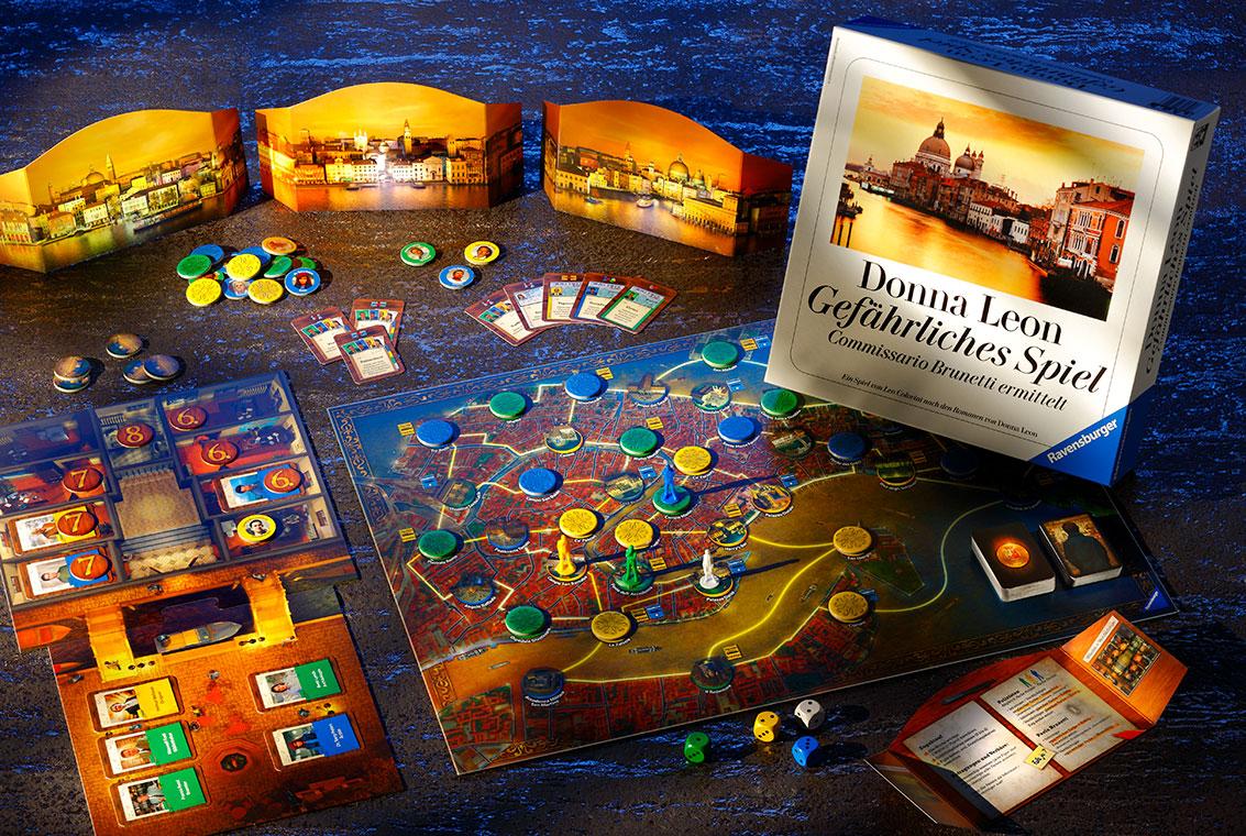 Ein Brettspiel liegt auf einem Tisch. Es zeigt eine farbenfrohe Spieloberfläche mit verschiedenen Spielfiguren, Karten und Spielmarkern. Neben dem Spielbrett steht eine Spieleschachtel mit dem Titel "Donna Leon - Gefährliches Spiel". Der Hintergrund zeigt eine stilisierte Stadtansicht.