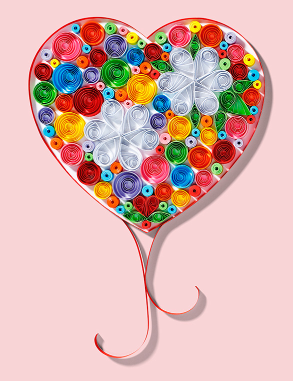 Ein herzförmiges Kunstwerk aus bunten Papierstreifen in unterschiedlichen Farben, verziert mit zwei weißen, blütenähnlichen Formen. Der Hintergrund ist rosa. Das Herz hat einen eleganten, geschwungenen Schwung am unteren Ende.