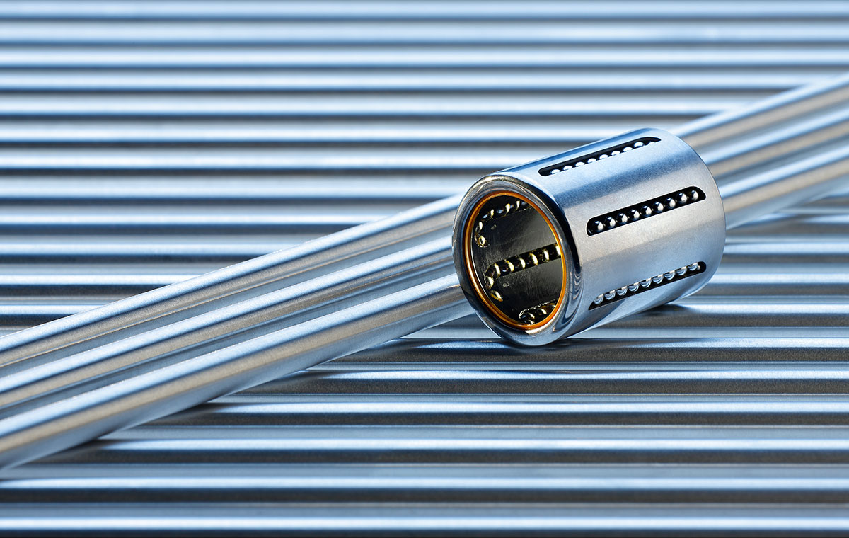 Ein Metallrohr liegt horizontal auf einer gestreiften Unterlage, während ein runder Metallzylinder darüber positioniert ist. Der Zylinder hat seitliche Öffnungen und einen glänzenden Korpus, was auf mechanische Funktionen hinweist.