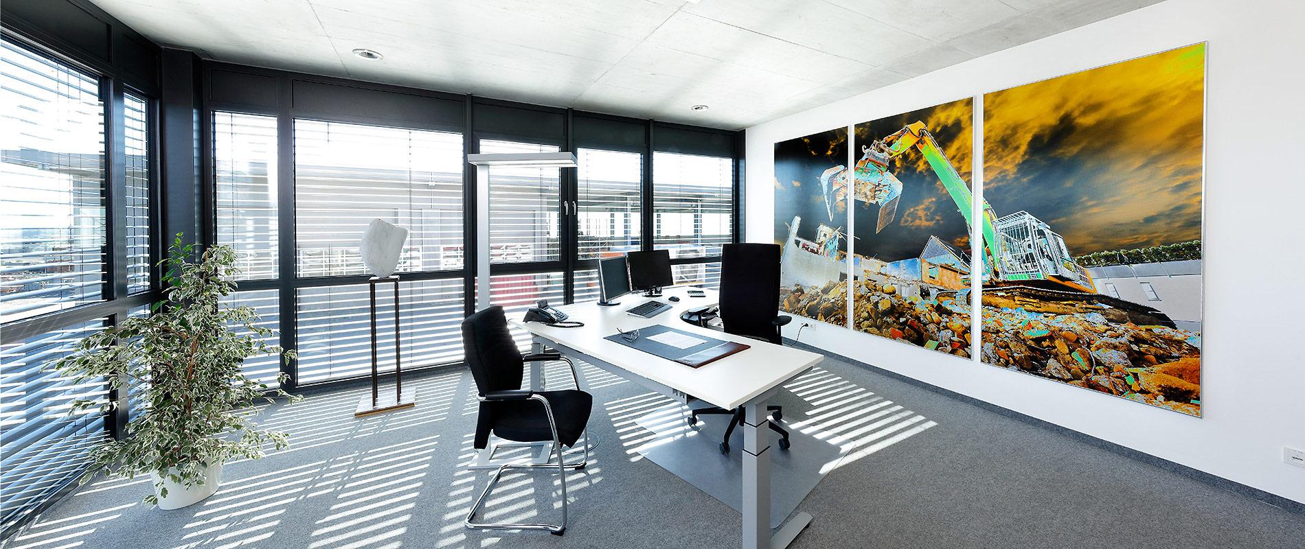 Grossformatdruck Ein modernes Büro mit großen Fenstern, die viel Tageslicht hereinlassen. Der Raum ist mit einem Schreibtisch, Stühlen und einer Pflanze eingerichtet. An der Wand hängt ein großes, buntes Bild, das eine Baustelle zeigt.
