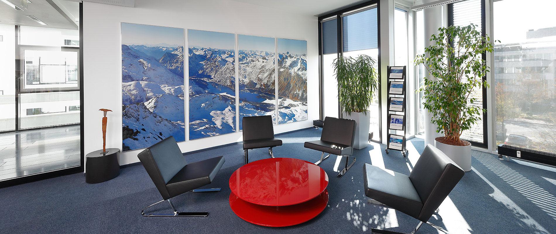 Grossformatdruck Moderne Wartebereich mit einem runden, roten Tisch in der Mitte und schwarzen Sesseln um ihn herum. Große Fenster lassen viel Licht herein, und an der Wand hängt ein großes Bild von einer Berglandschaft. Eine Pflanze steht ebenfalls im Raum.