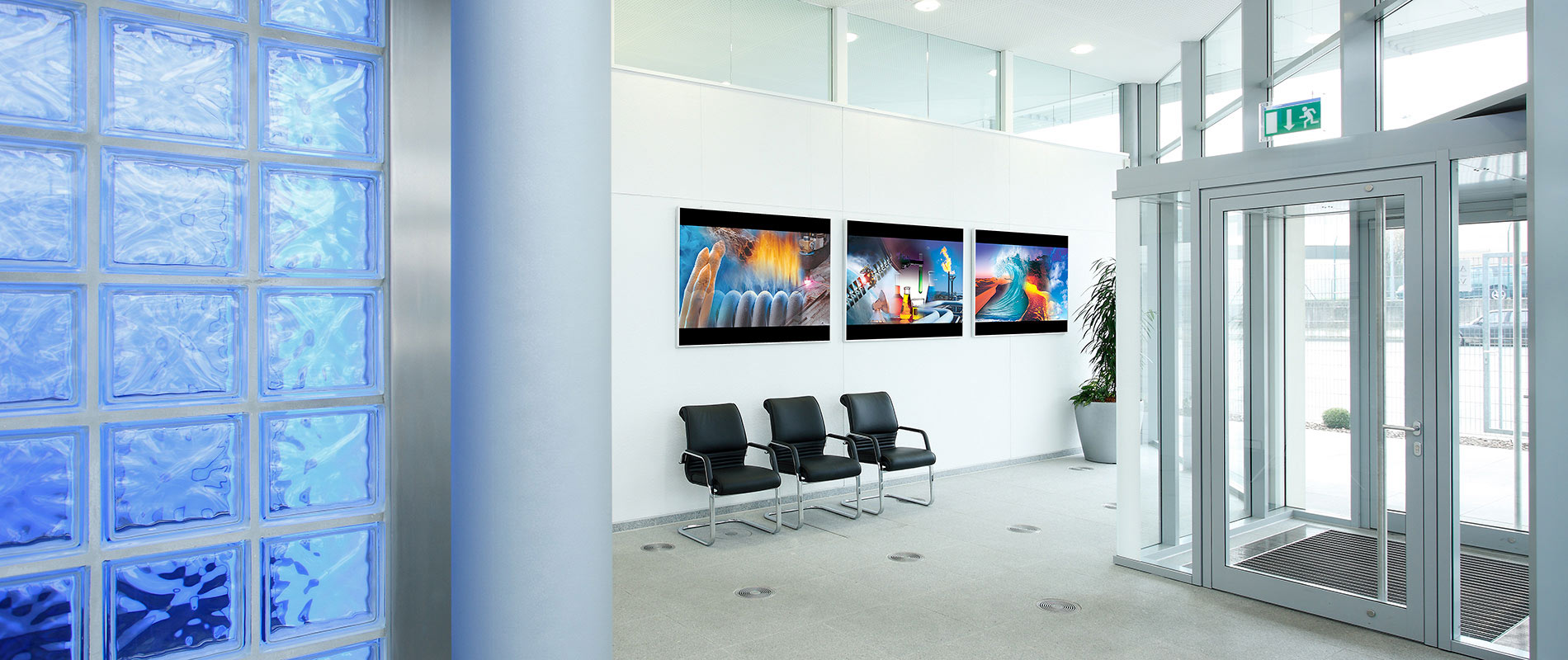 Grossformatdruck Ein moderner Empfangsbereich mit großen Fenstern und hellen Wänden. Auf einer Wand hängen drei Bilder, während auf dem Boden zwei schwarze Stühle stehen. Links ist eine Glassteinwand sichtbar, die durch das Licht ein attraktives Muster erzeugt.