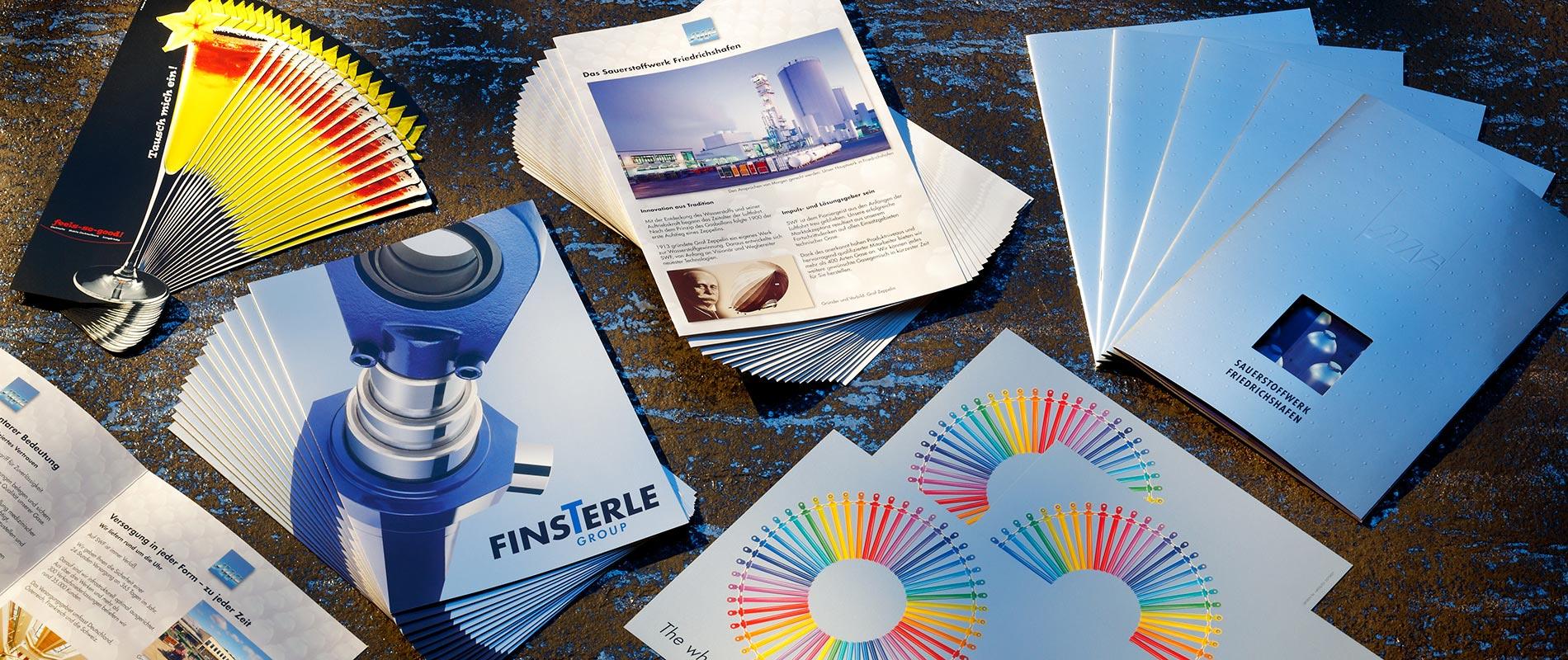 Drucksachen Eine Sammlung von Broschüren und Farbmustern auf einem Holzuntergrund. Die Werke zeigen verschiedene Designs, darunter Produkte und Informationen über das Unternehmen. Farbenfrohe Fächer und informative Dokumente sind sichtbar.