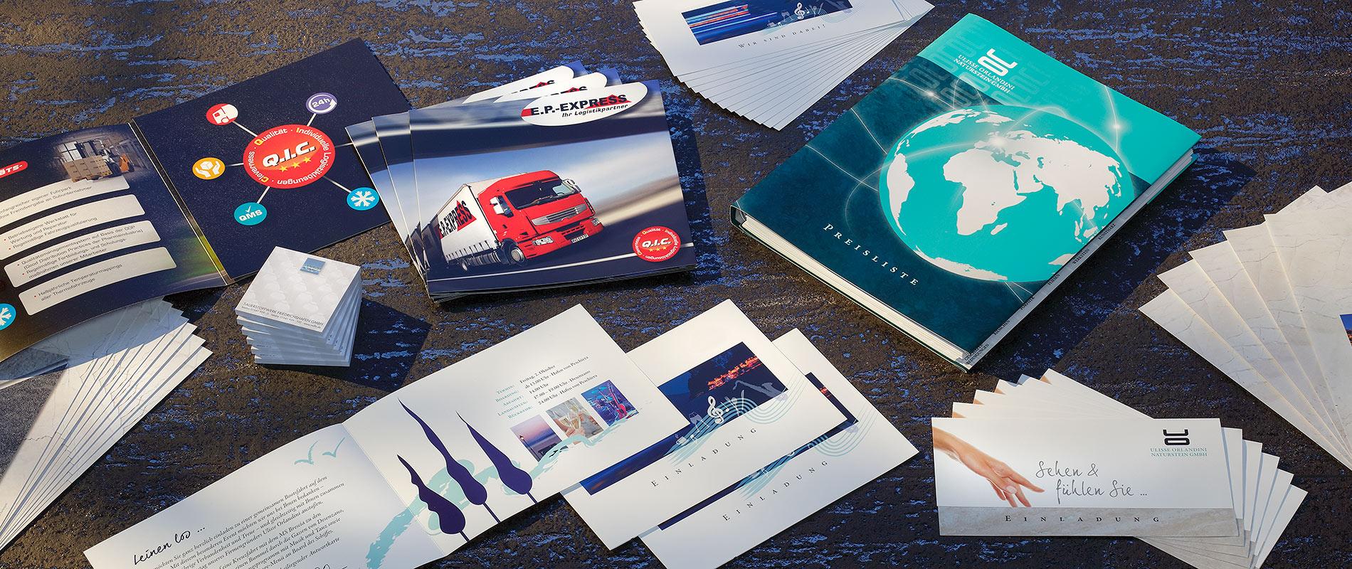 Drucksachen Eine Vielzahl von gedruckten Materialien liegt auf einem Tisch. Dazu gehören Broschüren, Flyer und Visitenkarten mit verschiedenen Designs, darunter Bilder von Lkw und einer Weltkarte. Die Farben sind überwiegend blau, rot und weiß.