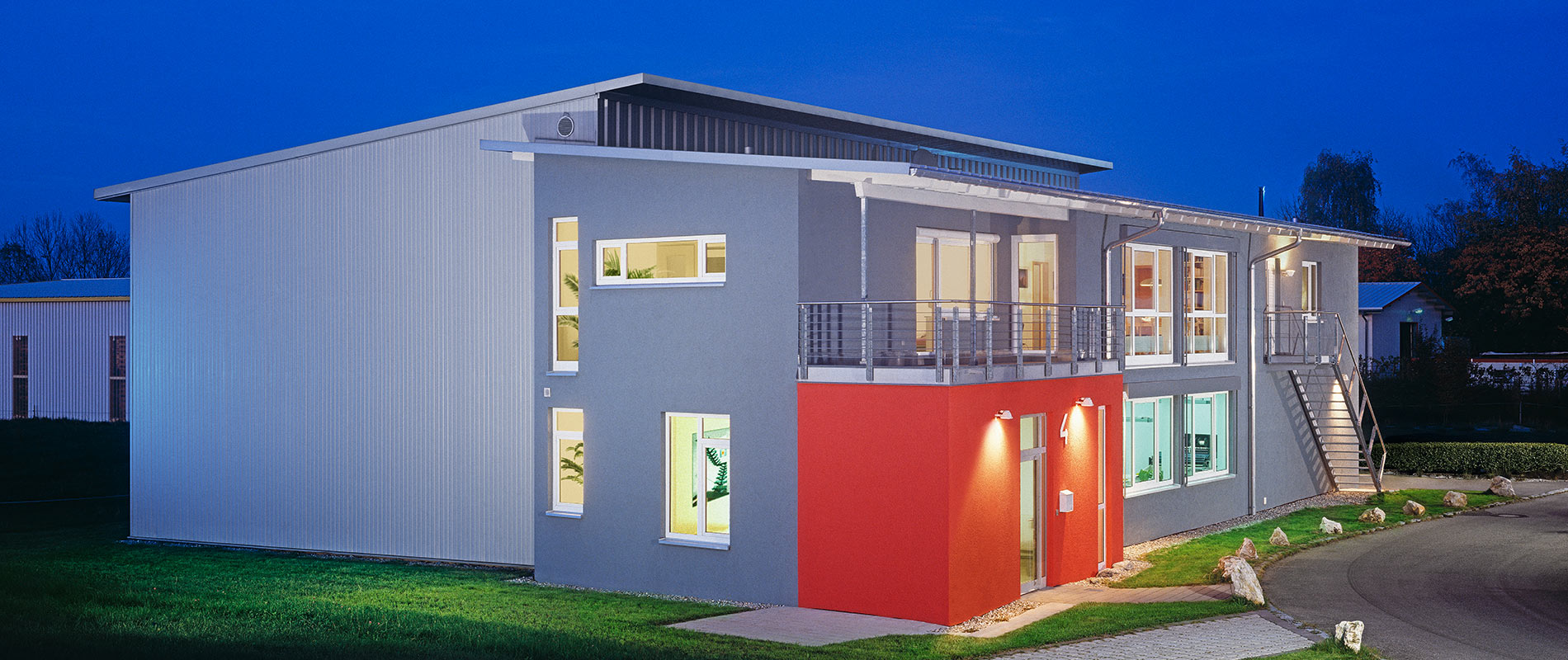 Moderne zweigeschossige Gebäudeansicht bei Nacht, mit grauer und roter Fassade. Licht strömt aus den Fenstern und schafft eine einladende Atmosphäre. Eine Treppe führt zum oberen Geschoss, umgeben von Grünflächen.