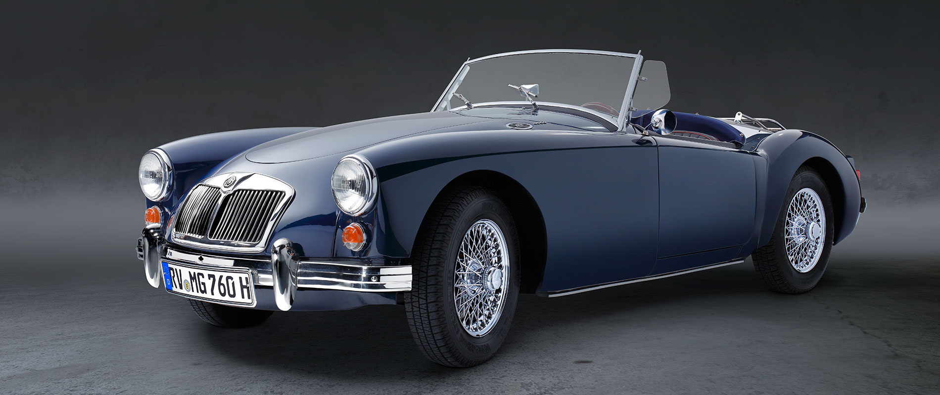 Ein klassischer blauer Sportwagen, ein Cabriolet mit offenem Verdeck. Der Wagen hat elegante Linien, Chromdetails und Drahtspeichenräder. Der Hintergrund ist dunkel, was den Wagen hervorhebt.