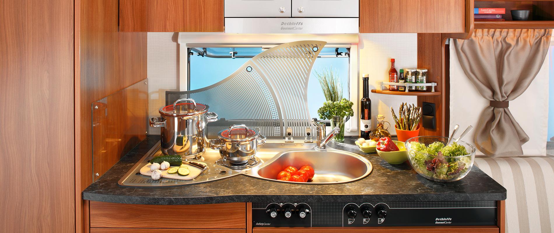 Moderne Küche mit Holzschränken und schwarzer Arbeitsplatte. Auf der Platte befinden sich frisches Gemüse, Kochutensilien und eine Spüle. Im Hintergrund sieht man ein Fenster, das einen Ausblick auf blauen Himmel und Pflanzen bietet.