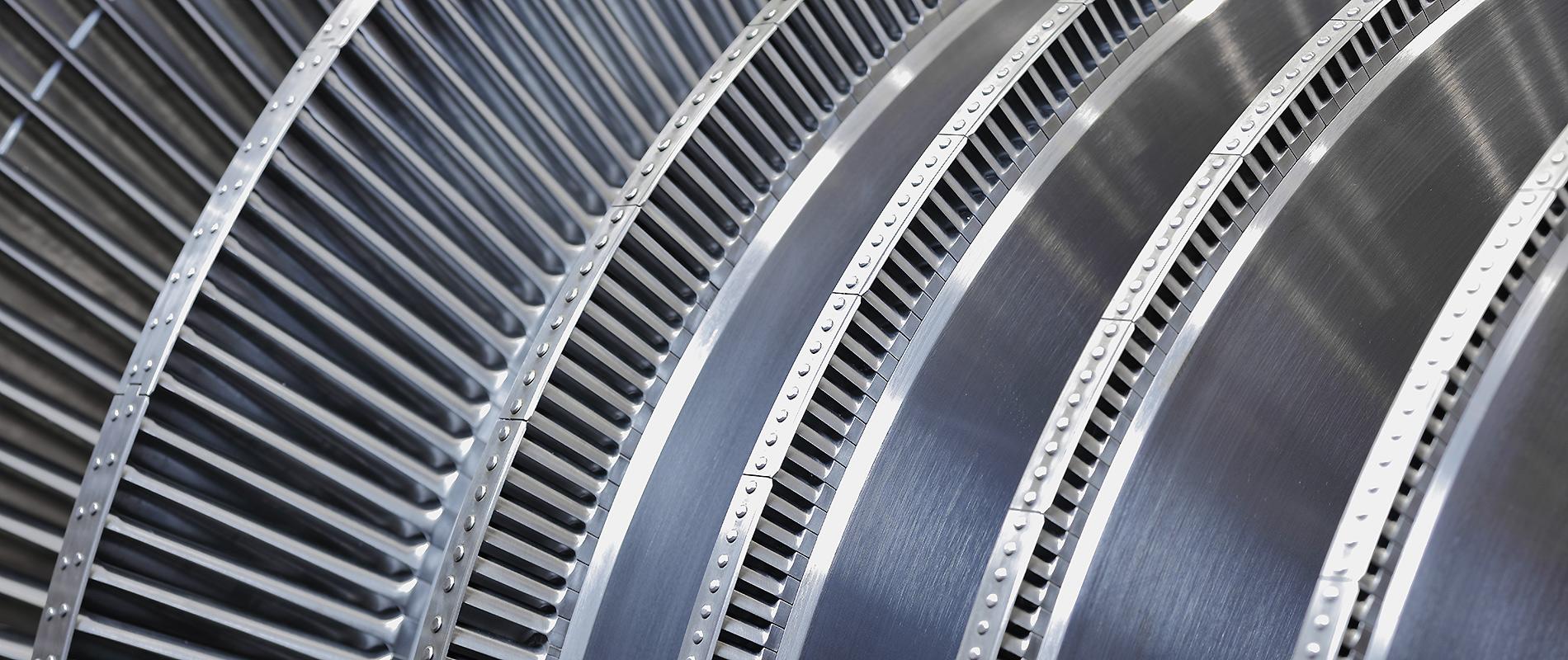 Nahaufnahme von mehreren Turbinenschaufeln aus Metall, die in einer geschwungenen Anordnung angeordnet sind. Die Oberfläche ist glänzend und zeigt präzise Bearbeitungslinien. Die Schaufeln sind eng nebeneinander angeordnet, was die technische Detailgenauigkeit betont.