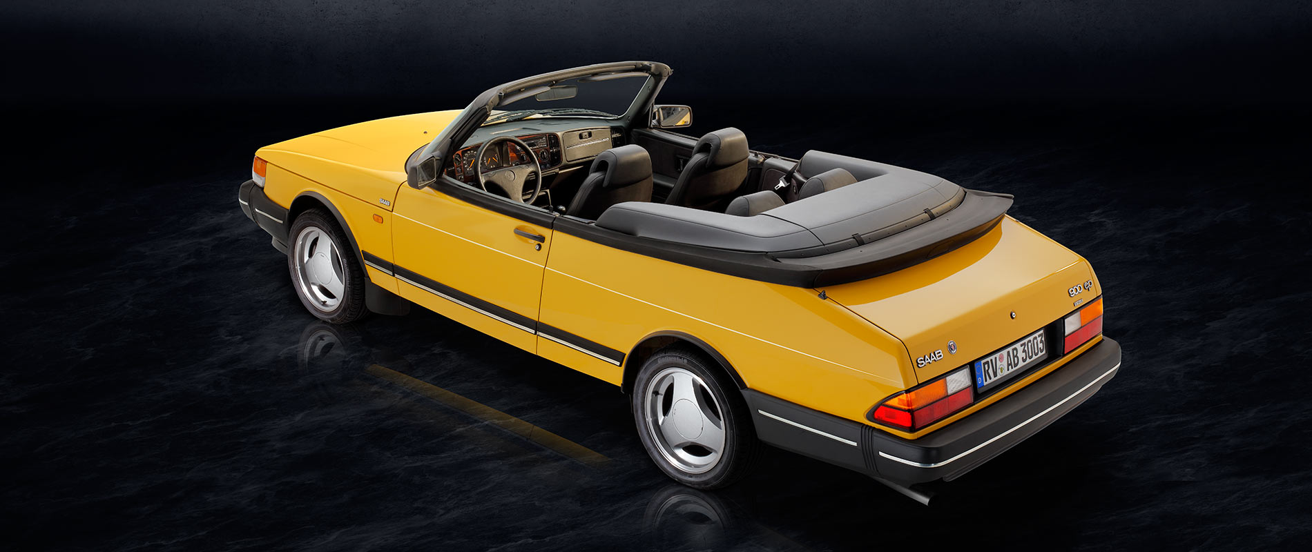 Ein gelb lackierter Cabriolet-Auto mit offenem Verdeck steht auf einem dunklen Hintergrund. Das Fahrzeug hat eine sportliche Form und elegante Felgen, während der Innenraum in dunklen Tönen gehalten ist.