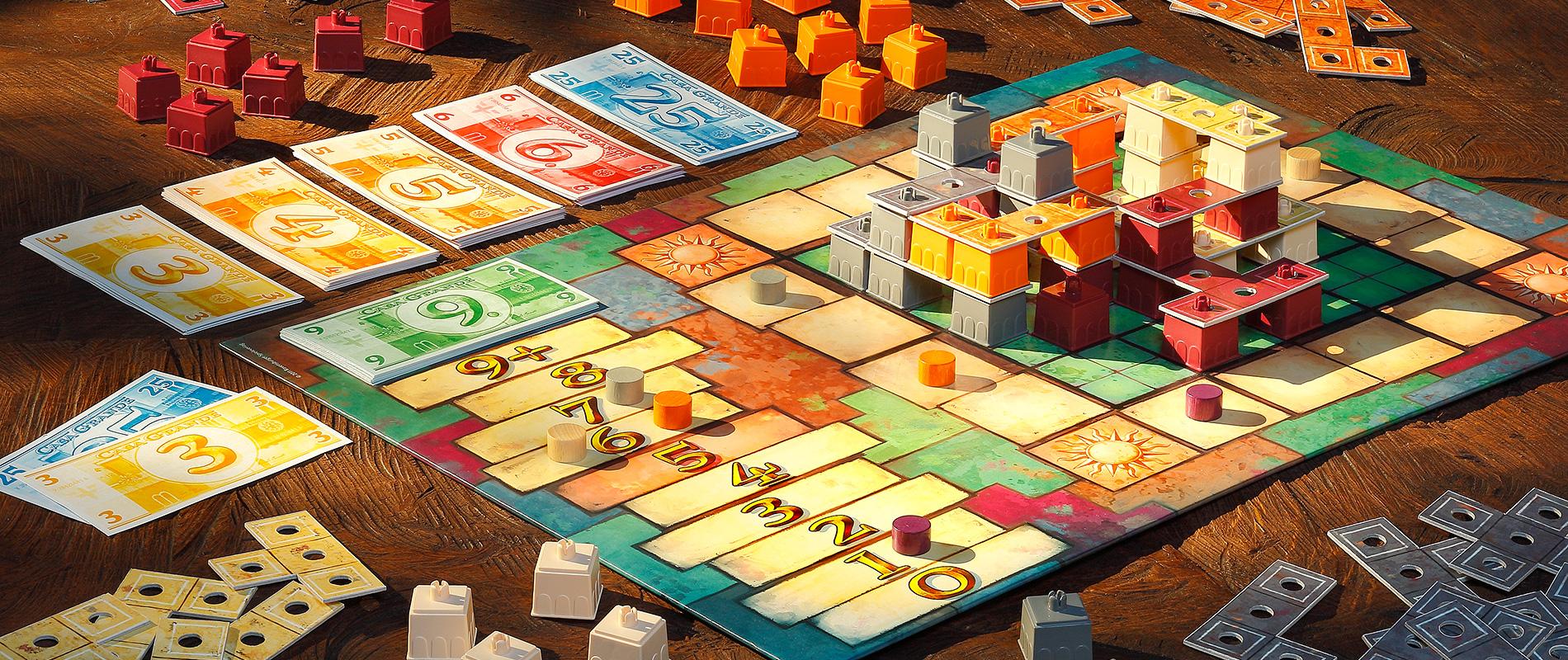 Ein farbenfrohes Brettspiel auf einem Holztisch. Das Spielbrett zeigt eine von bunten Spielsteinen aufgebaute Struktur in der Mitte. Daneben liegen Stapel mit Spielkarten und Münzen in verschiedenen Farben.