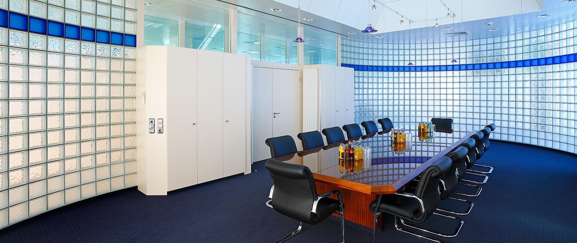 Ein moderner Besprechungsraum mit einem langen Holztisch, umgeben von schwarzen Stühlen. Die Wände sind mit Glasblock-Elementen gestaltet, und es gibt einige Schränke. Der Raum hat eine helle, offene Atmosphäre durch große Fenster.