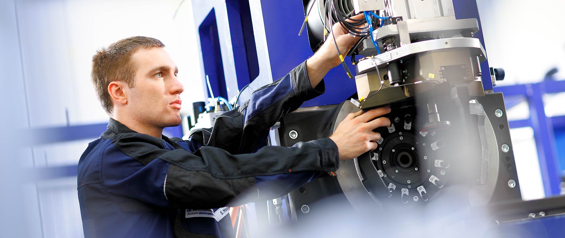 DL382 Ein Techniker arbeitet an einer Maschine, während er Kabel und Teile überprüft. Er trägt eine Arbeitsjacke und konzentriert sich auf die Installation oder Wartung der technischen Komponenten. Der Hintergrund zeigt industrielle Ausrüstung und Maschinen.