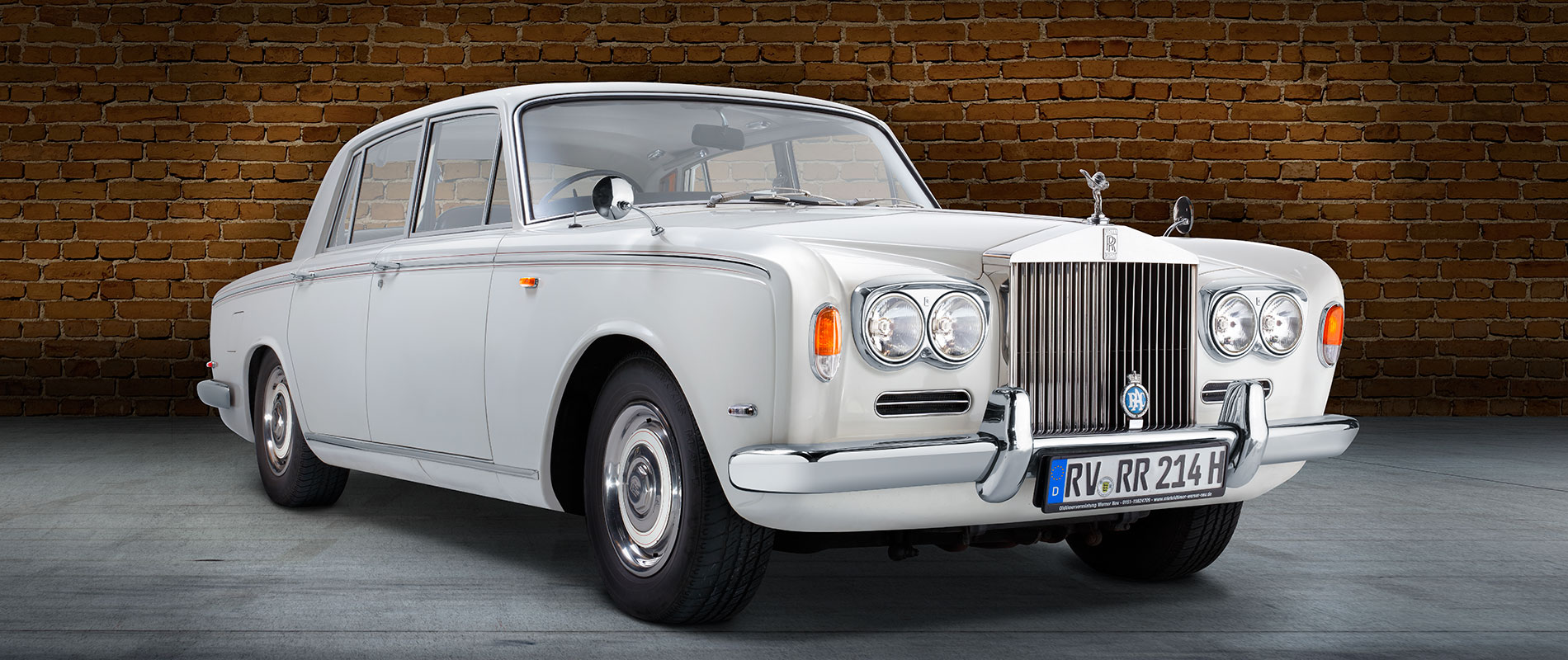 Rolls Royce Silver Shadow Front Ein eleganter, klassischer Rolls-Royce in Weiß steht vor einer Backsteinmauer. Der Luxuswagen hat vier Türen, runde Scheinwerfer und ein markantes Kühlergrill mit dem Rolls-Royce-Emblem. Der Hintergrund besteht aus einer stilvollen grauen Fläche.