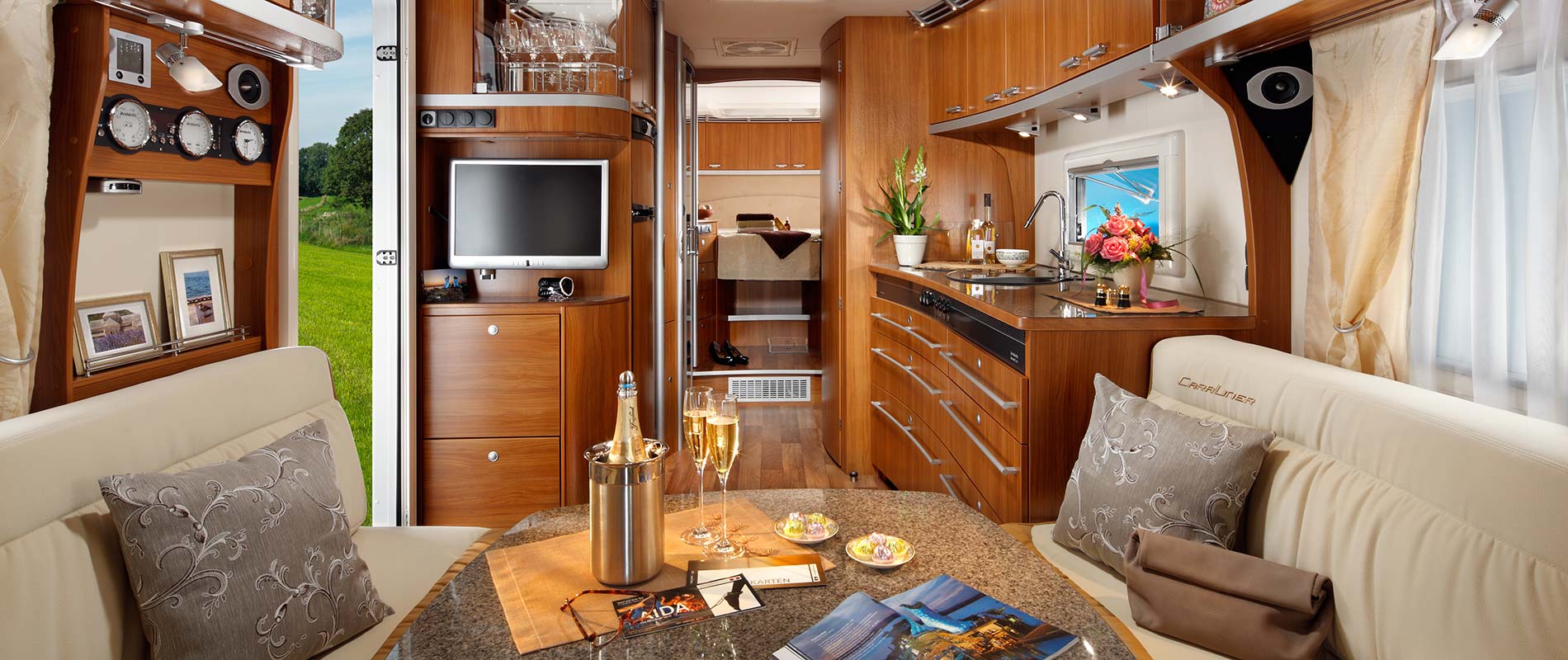 Luxuswohnwagen Eine modern gestaltete Innenansicht eines Wohnmobils mit holzverkleideten Wänden. Die Küche ist gut ausgestattet mit Geräten, während ein gemütlicher Sitzbereich mit einem Tisch und Dekorationen zu sehen ist. Fenster bieten einen Blick ins Grüne.