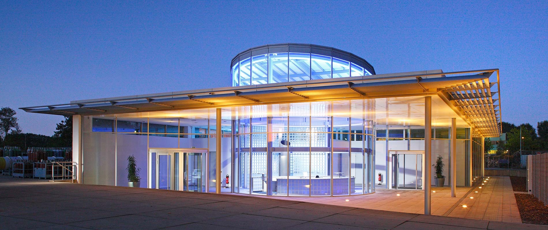 Empfangsgebaeude Moderne Architektur eines Gebäudes bei Dämmerung, das große Fensterfronten und einen runden Glaskuppelbereich aufweist. Das Gebäude ist beleuchtet und verfügt über einen großzügigen Eingangsbereich mit einer freundlichen Atmosphäre. Pflanzen sind in der Umgebung sichtbar.