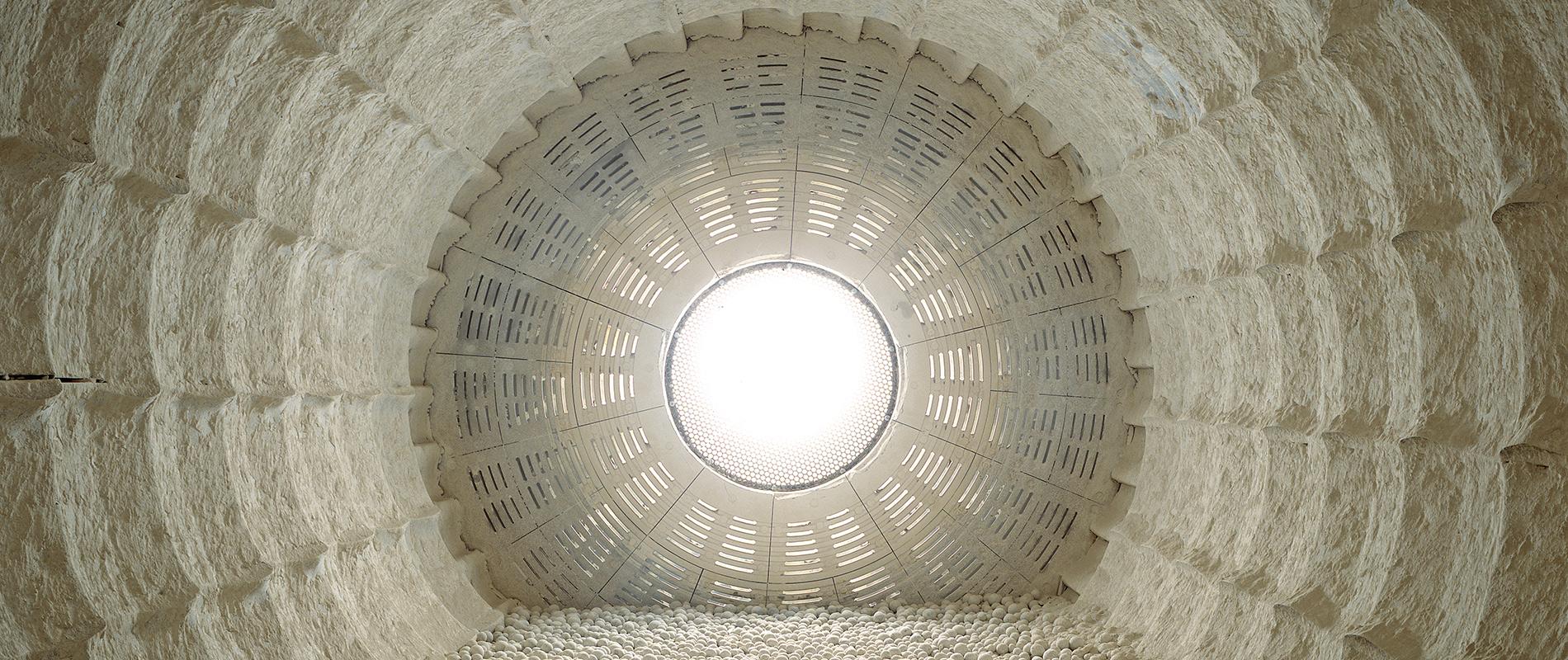 Innenansicht einer Rohmuehle Blick von innen nach oben durch eine runde, strukturierte Öffnung, die von hell erleuchtetem Licht gefüllt ist. Die Wände umgeben den Betrachter mit einem Muster aus regelmäßig angeordneten, vertikalen und horizontalen Linien.